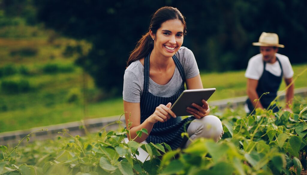 A woman smiles at the camera. She's crouching and holding a tablet.